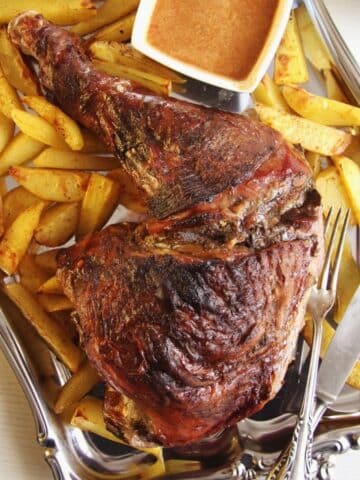 large slow roasted turkey leg served with roasted potatoes and a bowl of gravy on a silver platter.