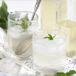two glasses with mint syrup drinks with ice cubes and bottles with syrup behind.