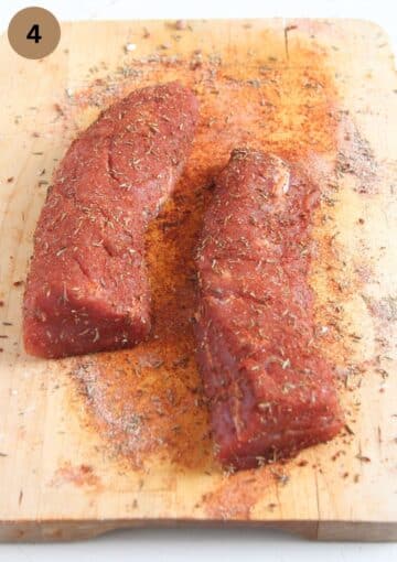 two raw pieces of pork generously seasoned on a wooden cutting board.