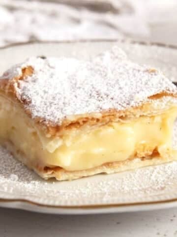 a slice of cremeschnitte with creamy vanilla filling and sprinkled with powdered sugar on a vintage plate.