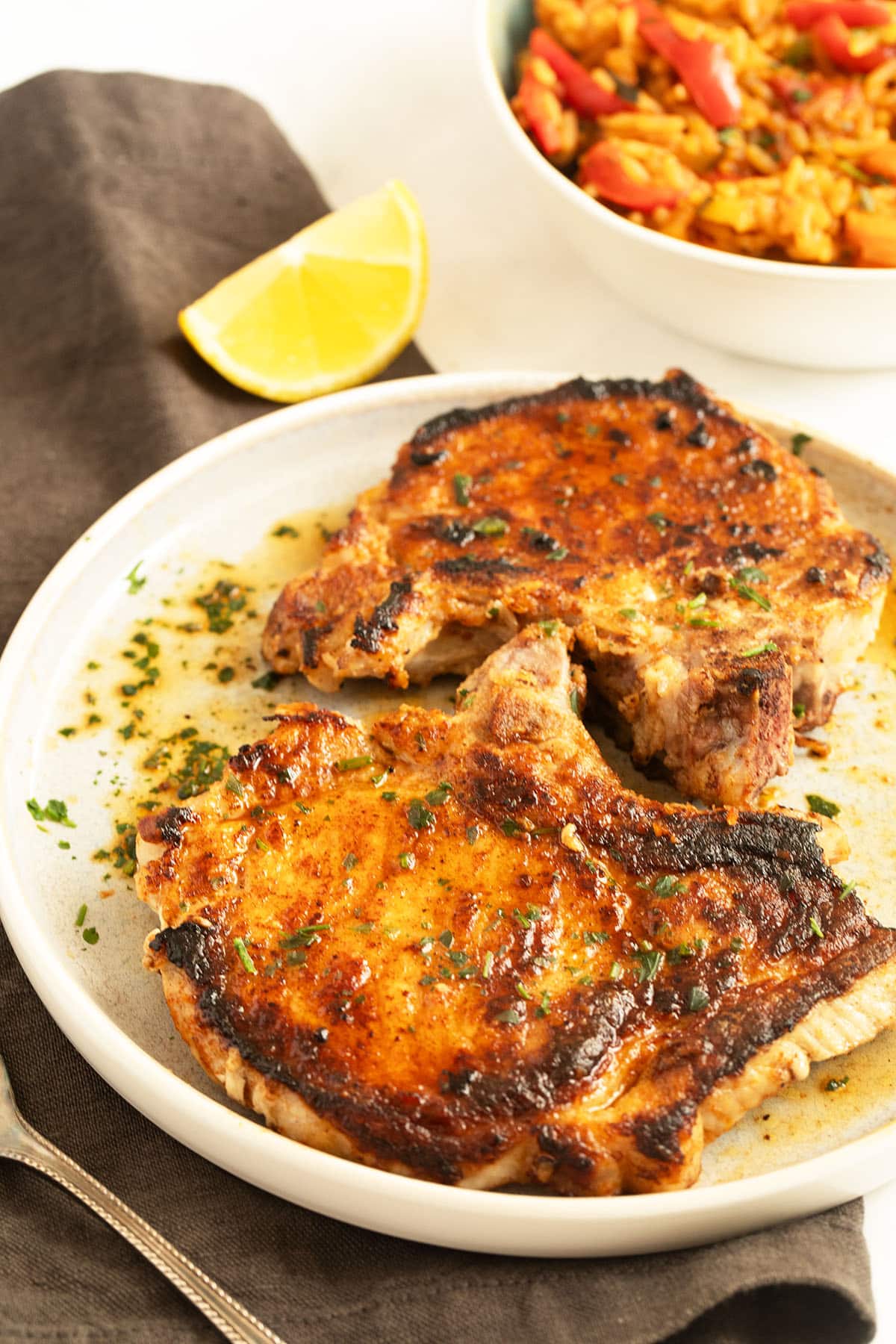 milk-brined pork chops with cooking juices on a plate, a bowl of rice and lemon behind it.
