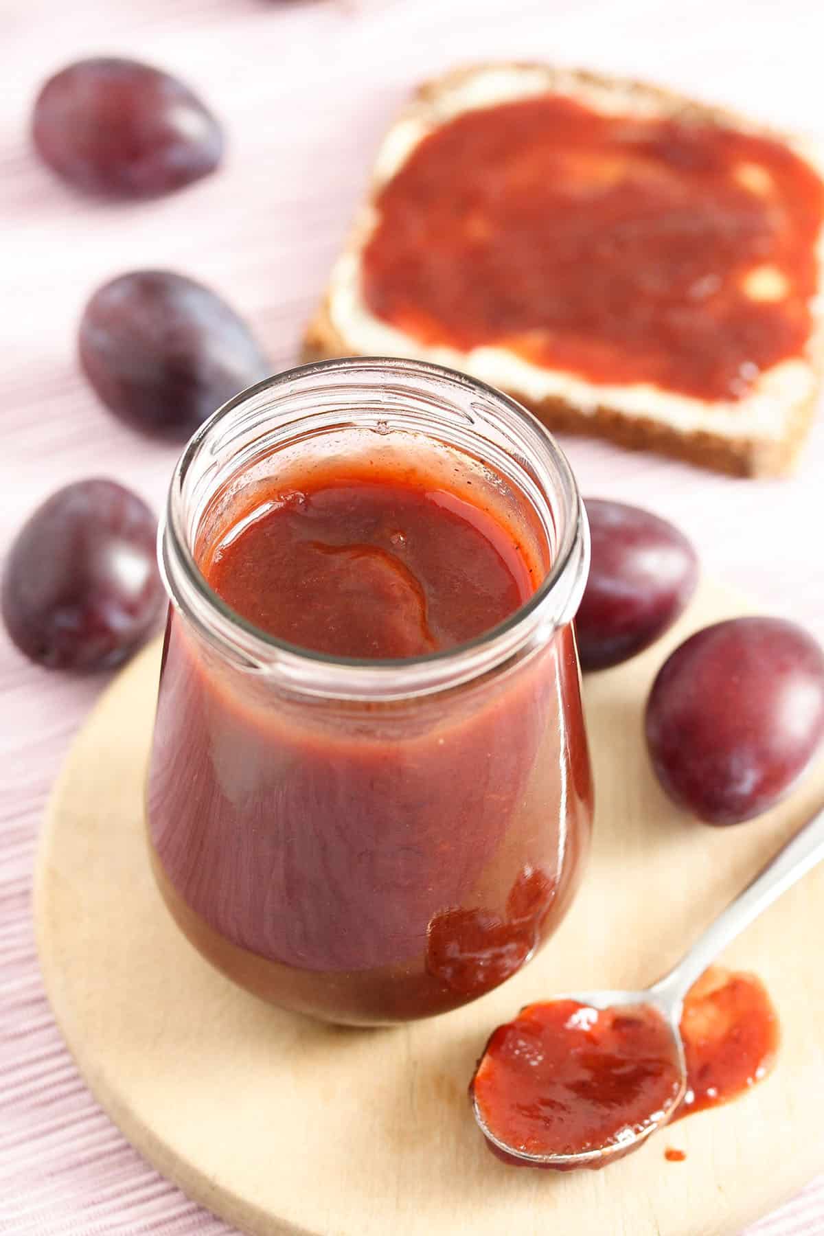 a small jar of german plum butter, many plums, a spoon and a slice of bread with butter and jam.