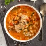 bowl of pork rib soup showing the ribs and the white beans on a grey kitchen cloth with a spoon beside it.