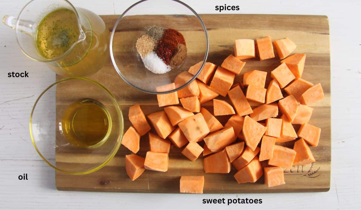 cubed raw sweet potatoes, vegetable broth, oil and spices on a wooden cutting board. 