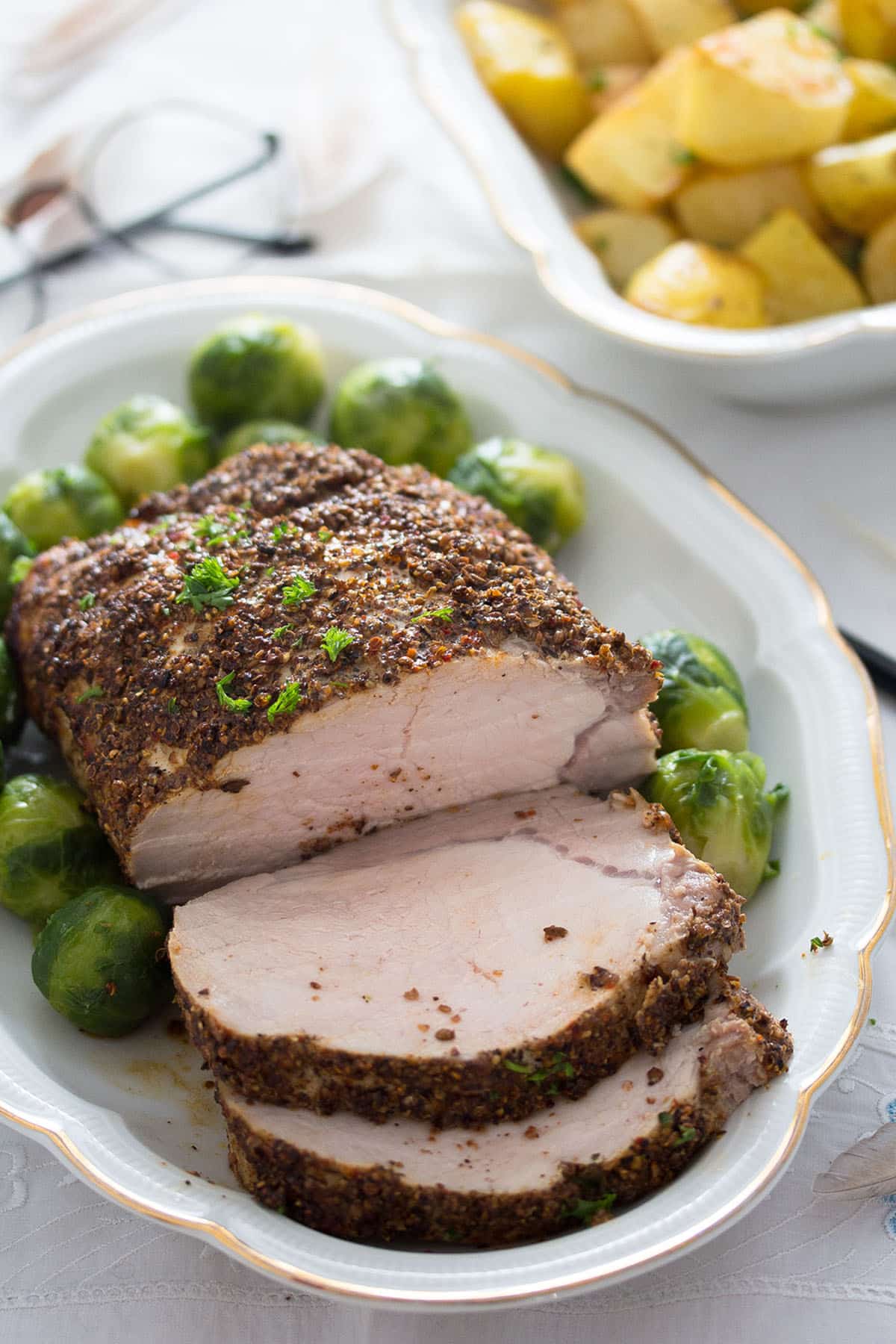 sliced pork roast with spicy crust on a platter, a bowl or potatoes behind.