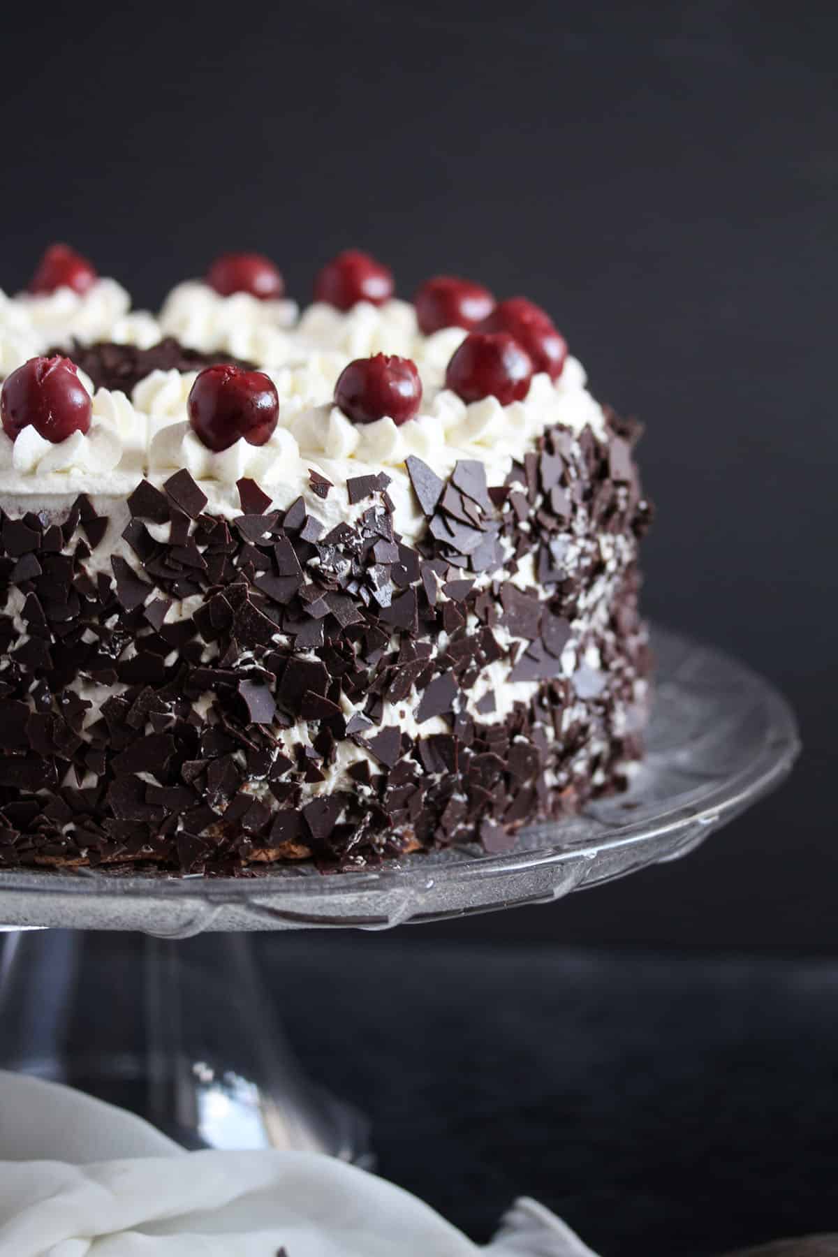 black forest cake decorated with chocolate shavings, cherries and cream on a high platter.