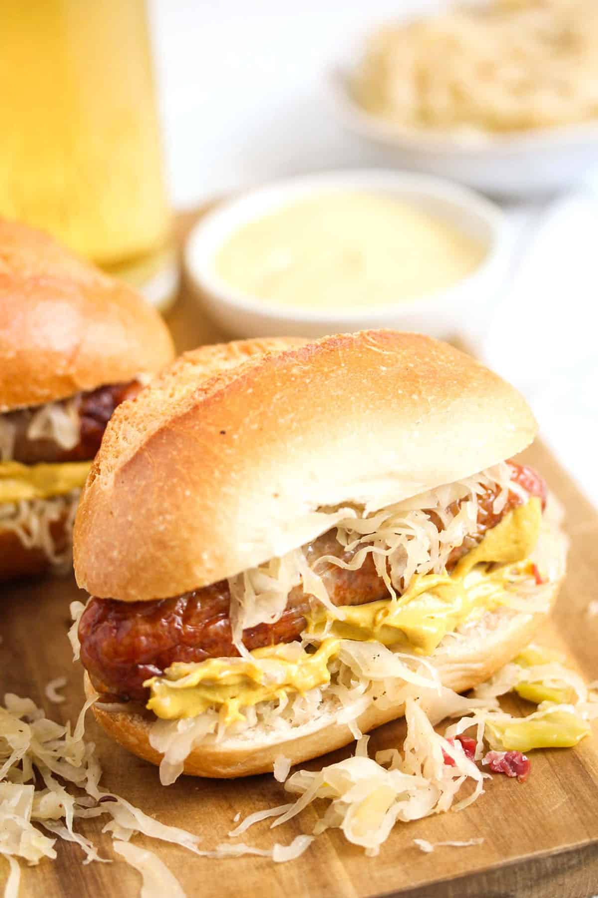 one german bun with bratwurst and sauerkraut, a small bowl of mustard and a beer behind.