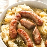 large bowl full with german kraut with whole and cut bratwurst on top. glass of water and potato mash behind.