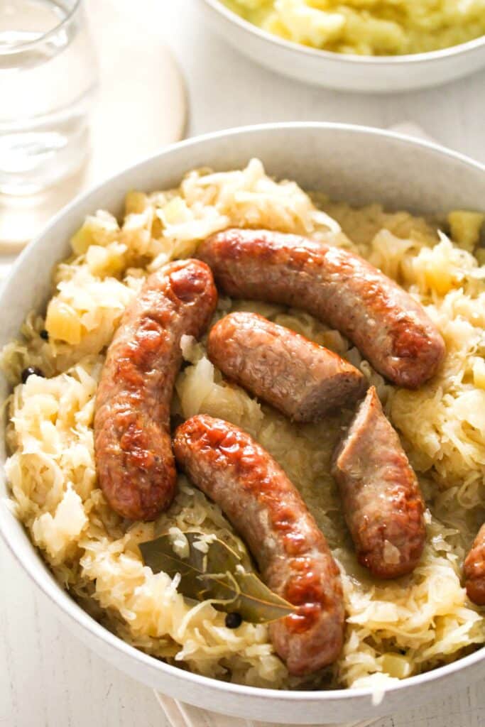 large bowl full with german kraut with whole and cut bratwurst on top. glass of water and potato mash behind.