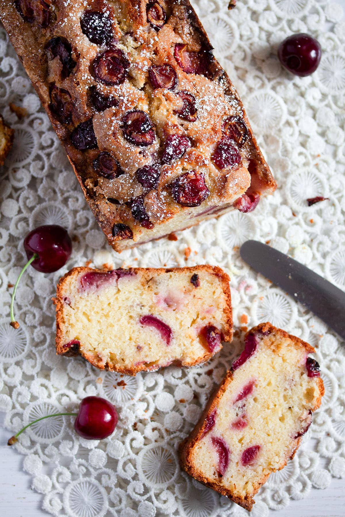 one cherry loaf cake, two cut slices, a small knife and a few fresh cherries.