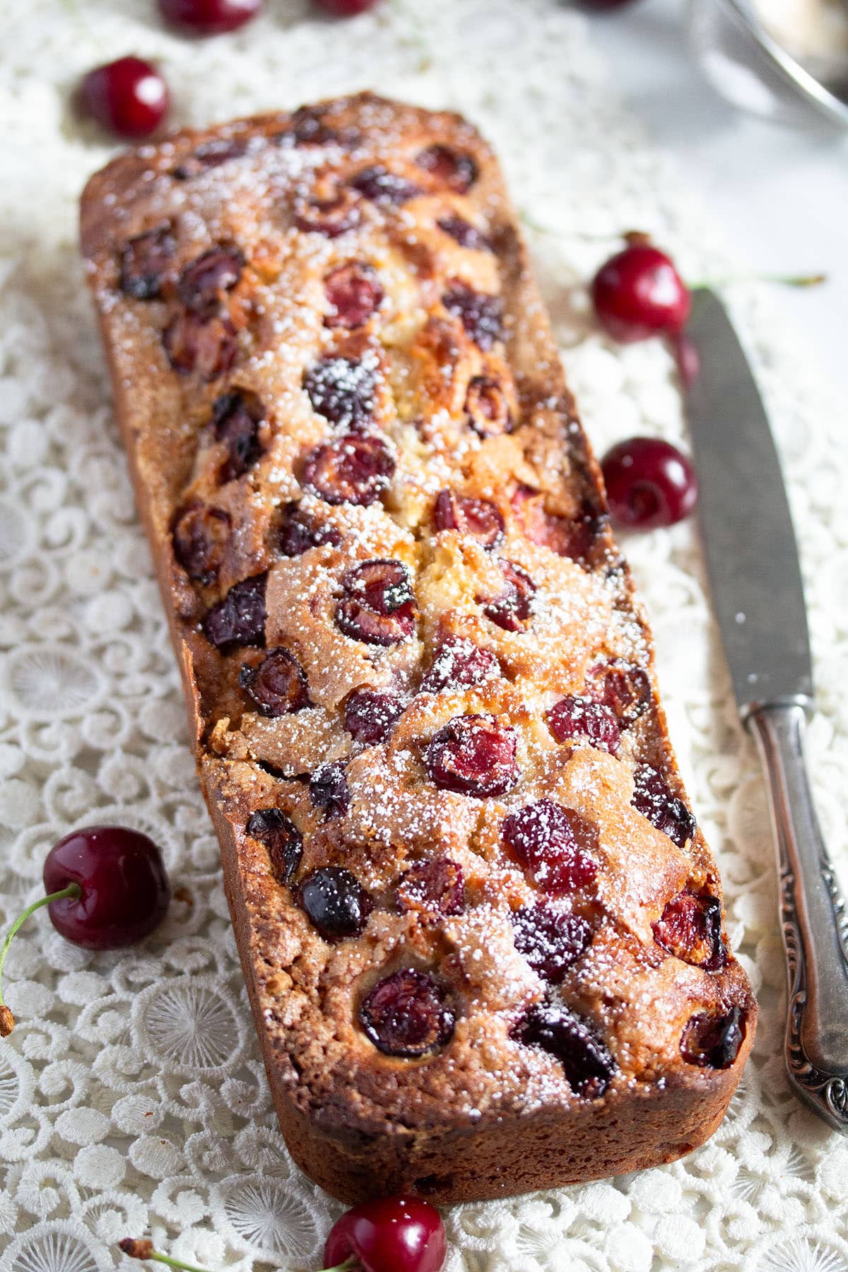 cherry loaf cake sprinkled with icing sugar, a knife, and some fresh cherries on the table.