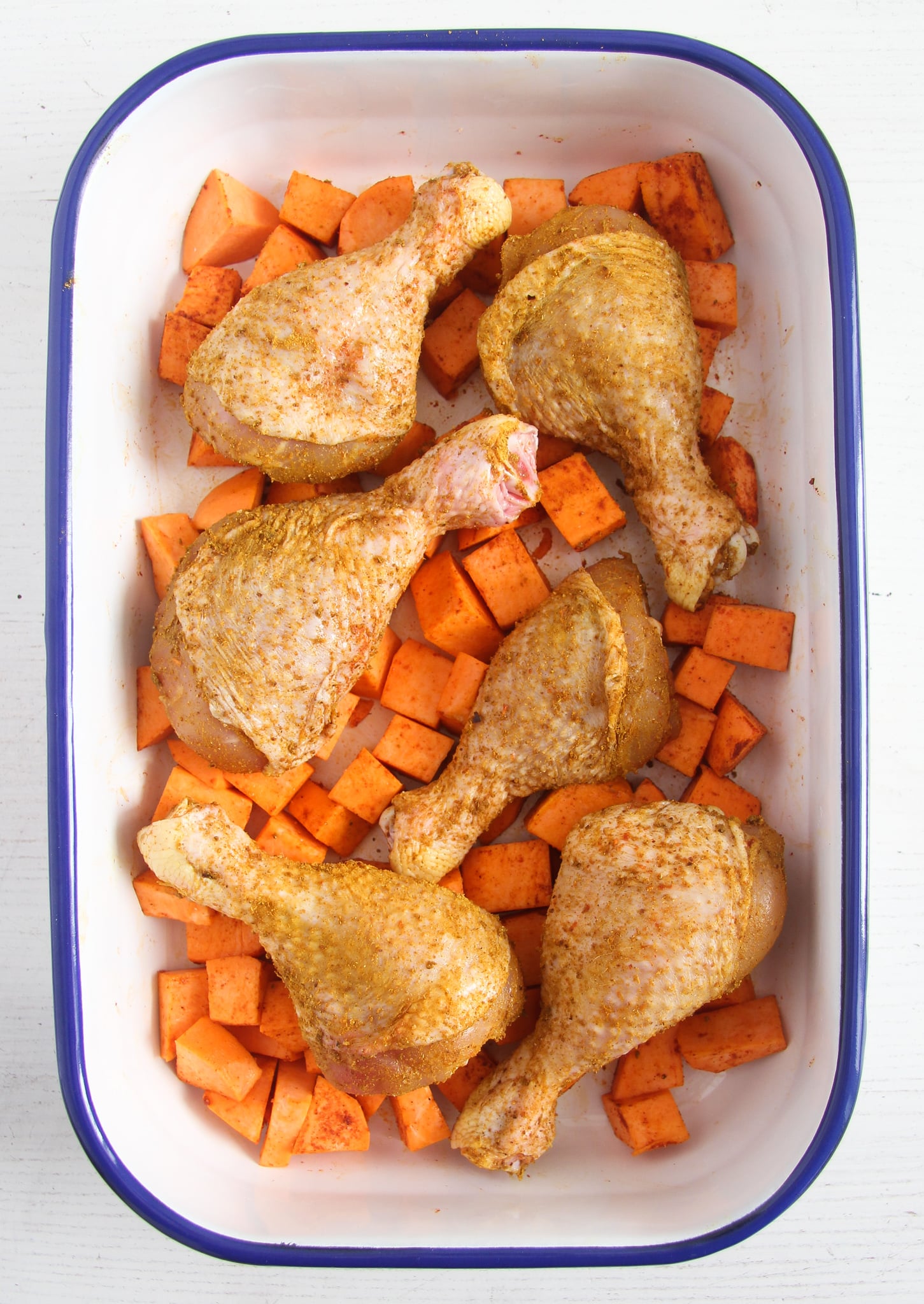 raw seasoned chicken legs and sweet potaotes in a white roasting tin with blue rim.