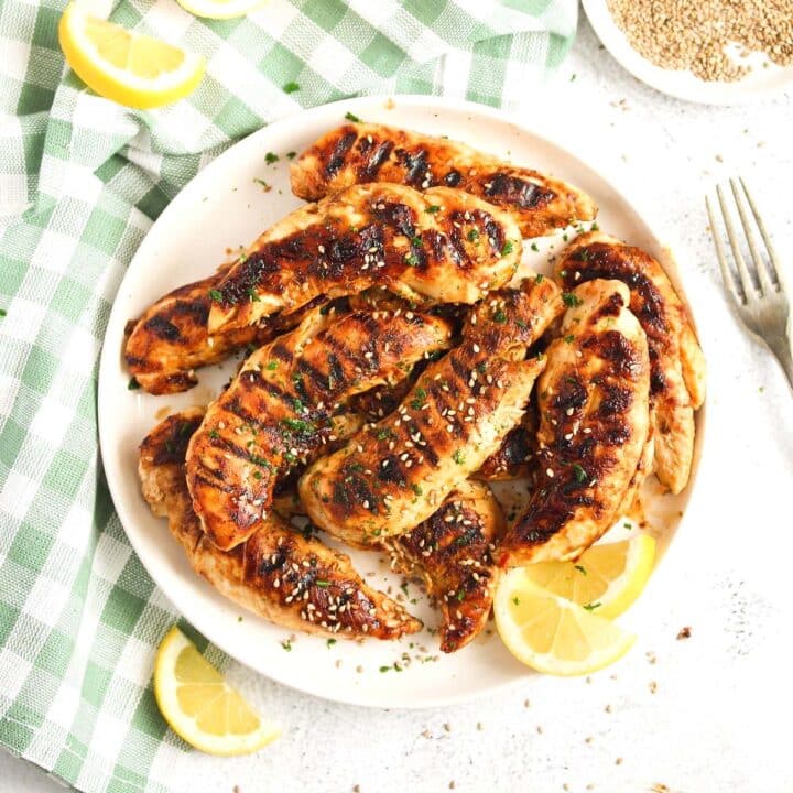 grilled chicken tenders served on a white plate with lemon wedges and sesame seeds.