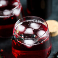 two glasses of diluted black currant syrup served with ice and a bottle behind.