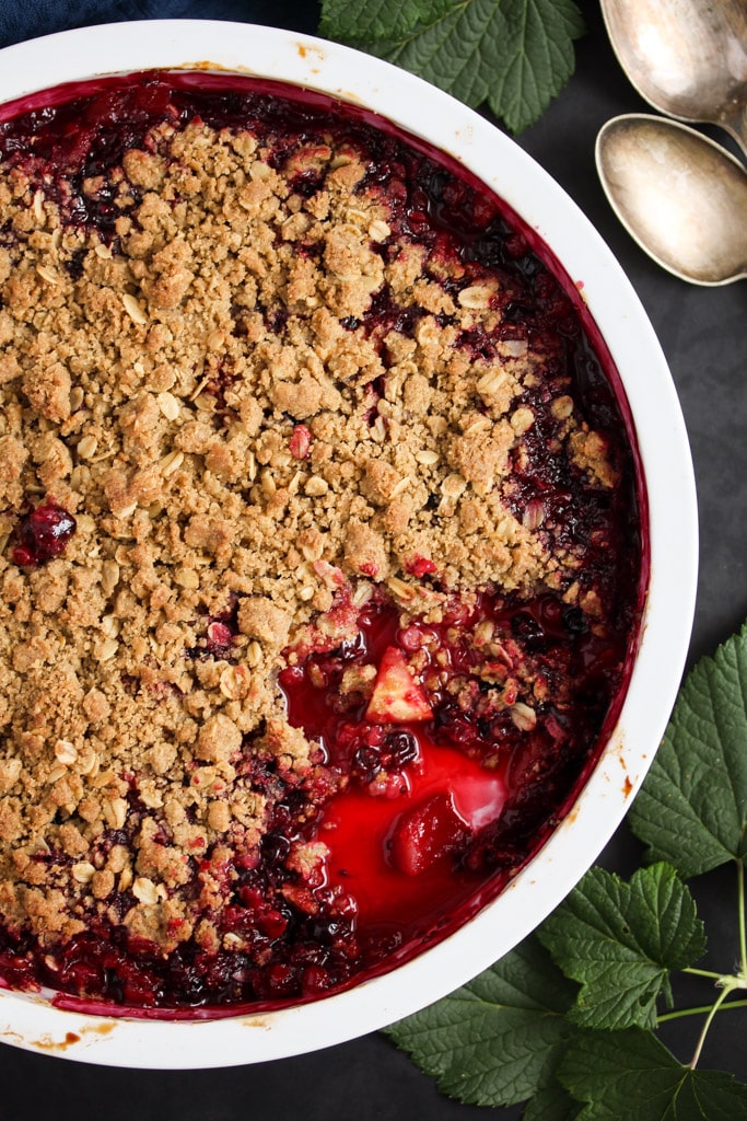 close up baking dish with currant crumble on a dark background, two tablespoons beside it.