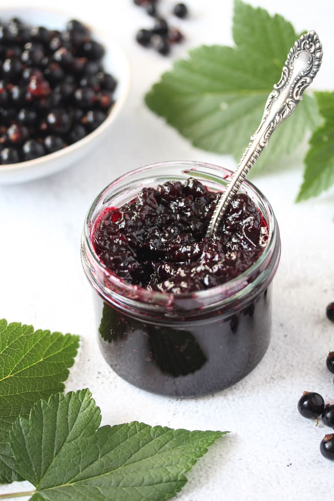 small jar of berry jam with a spoon in it and many leaves around it.