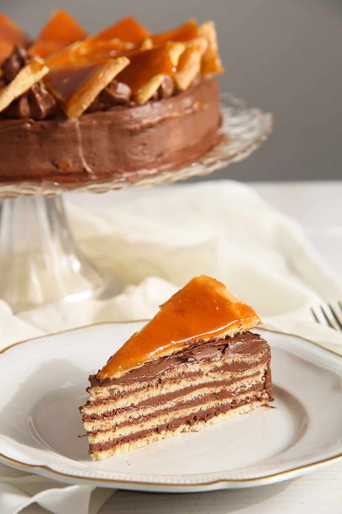 one slice of dobos torte in front and the rest of teh cake behind it on a tall platter.