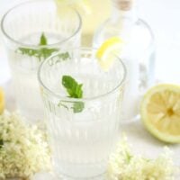 elderflower gin cocktail in two glasses, elderflowers and lemon around it.