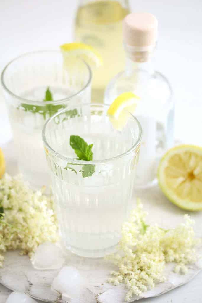 elderflower gin cocktail in two glasses, elderflowers and lemon around it.