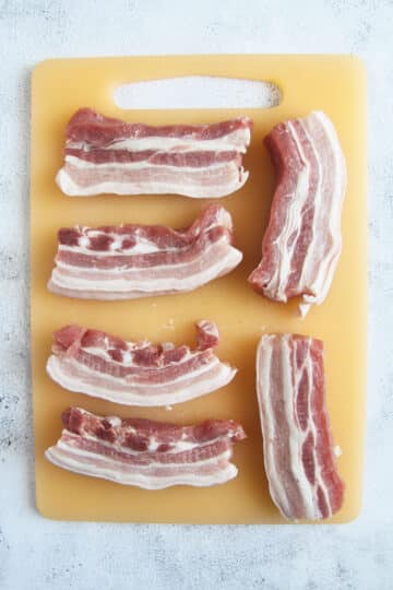 six raw pork belly strips on a cutting board.