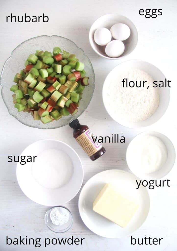 bowls of chopped rhubarb, three eggs, vanilla, sugar, flour, yogurt, butter, baking powder on a table.