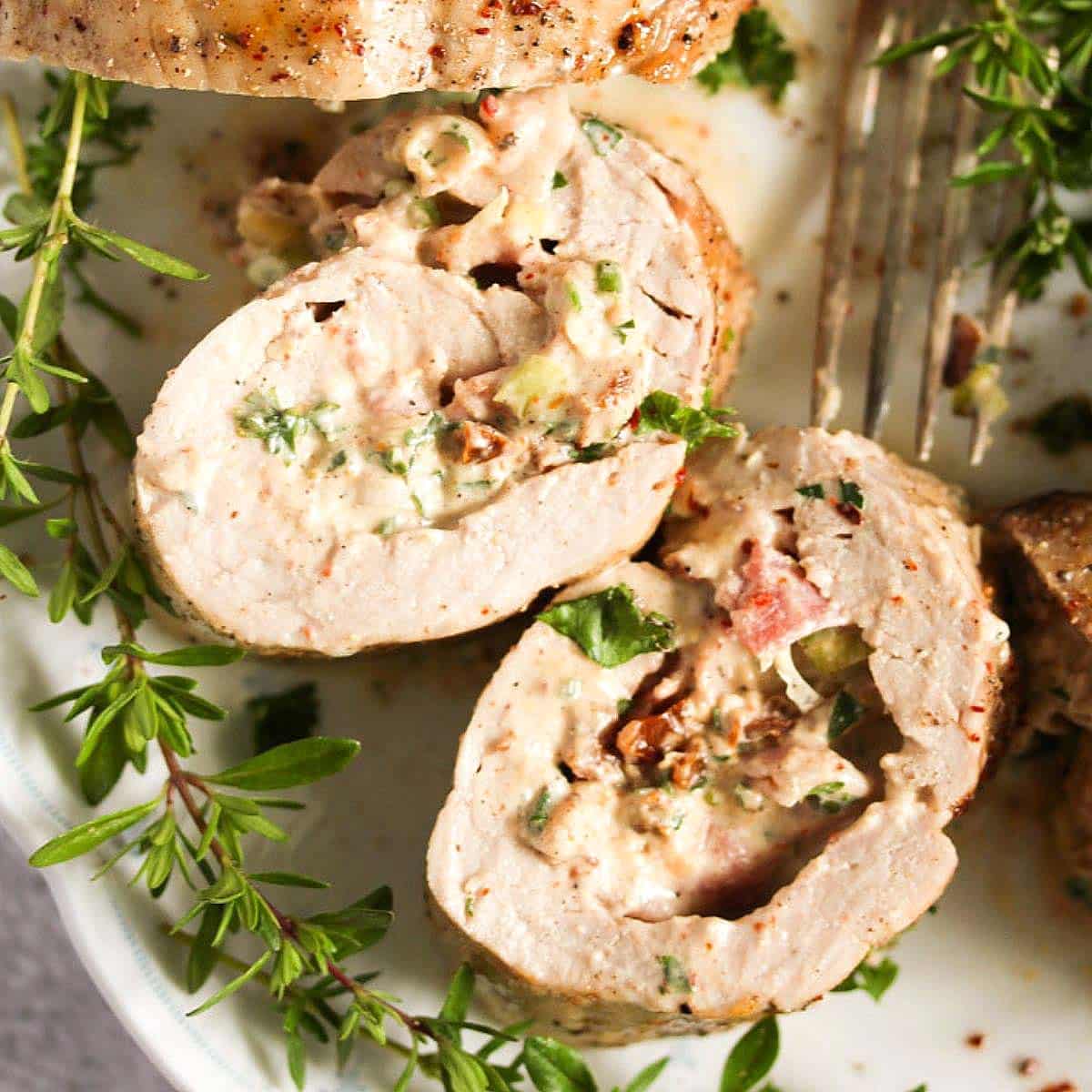 Two slices of cream cheese stuffed pork tenderloin with bacon on a platter with fresh herbs.