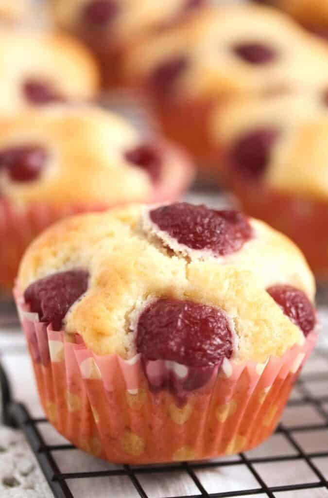 close up of a plump golden muffin topped with cherries on a wire rack.