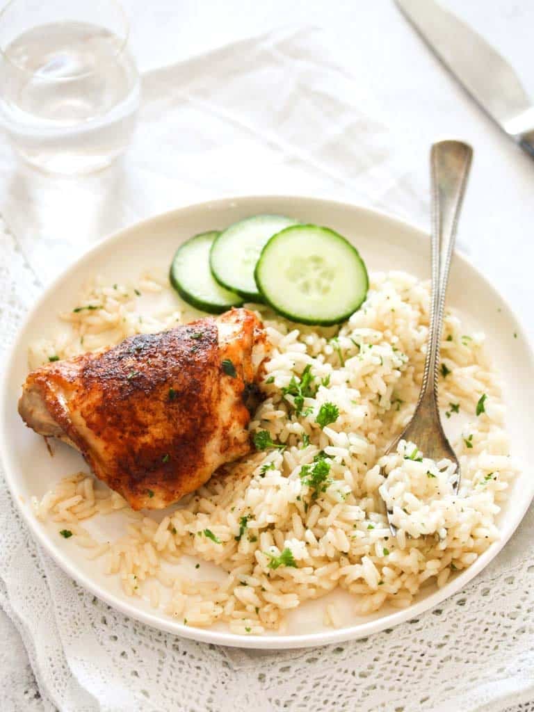 serving rice and chicken with cucumber on a small white plate with a fork and a glass behind.