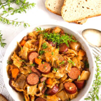 overhead view of cabbage with sausages in a white bowl with bread and thyme around it.