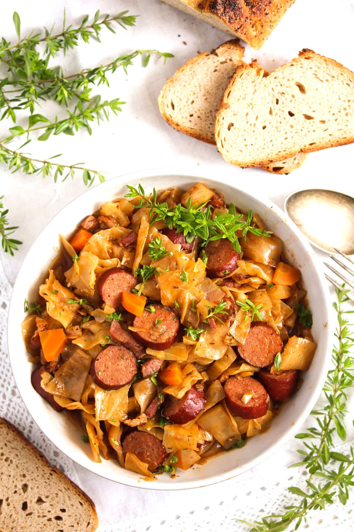 overhead view of cabbage with sausages in a white bowl with bread and thyme around it.