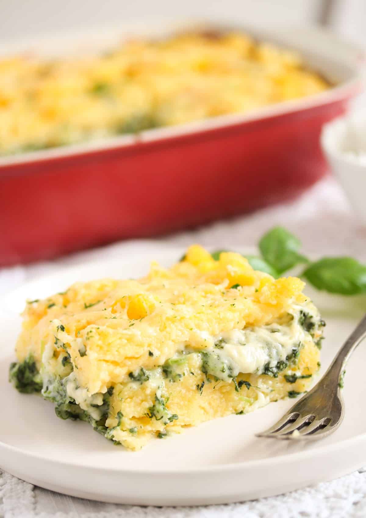 slice of polenta and spinach bake with a red casserole dish behind it.