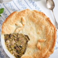 overhead view of chicken, mushrooms and leek pie with puff pastry on a blue white cloth with a spoon.