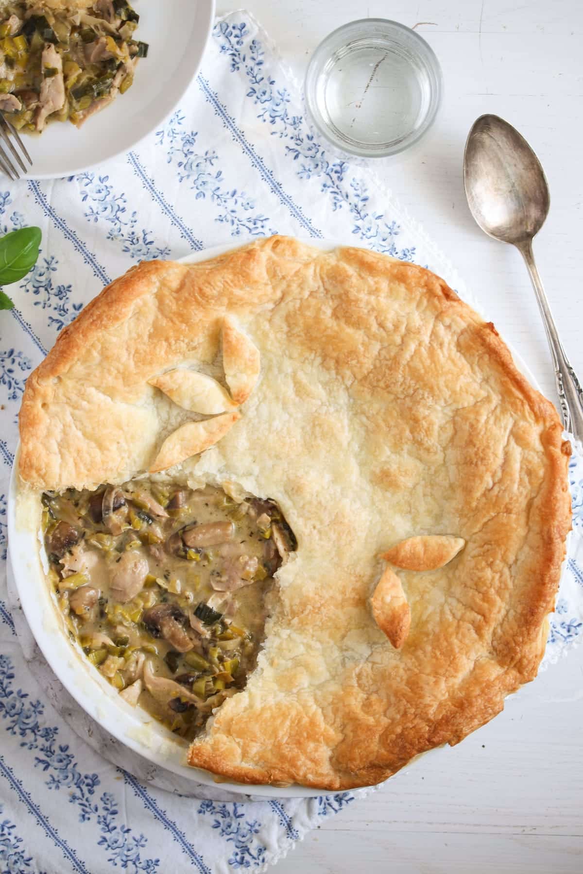 overhead view of chicken, mushrooms and leek pie with puff pastry on a blue white cloth with a spoon.