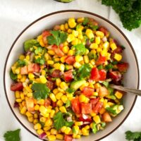 overhead view of a bowl with corn, tomatoes and herbs, half an avocado beside it.