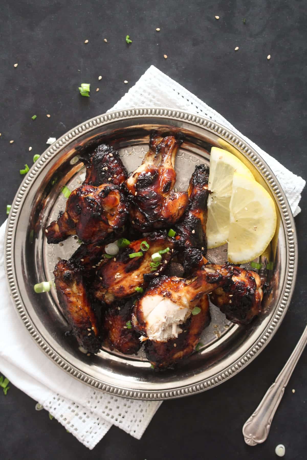 overhead view of a plate of dark coated chicken wings with lemon wedges.