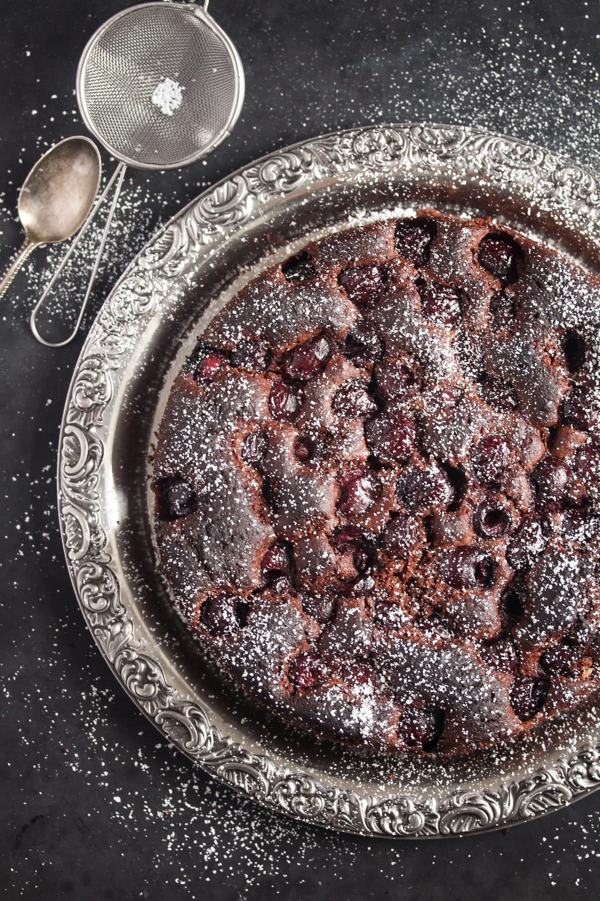 overhead view of cherry cake with chocolate on a silver plate with a small sieve beside.