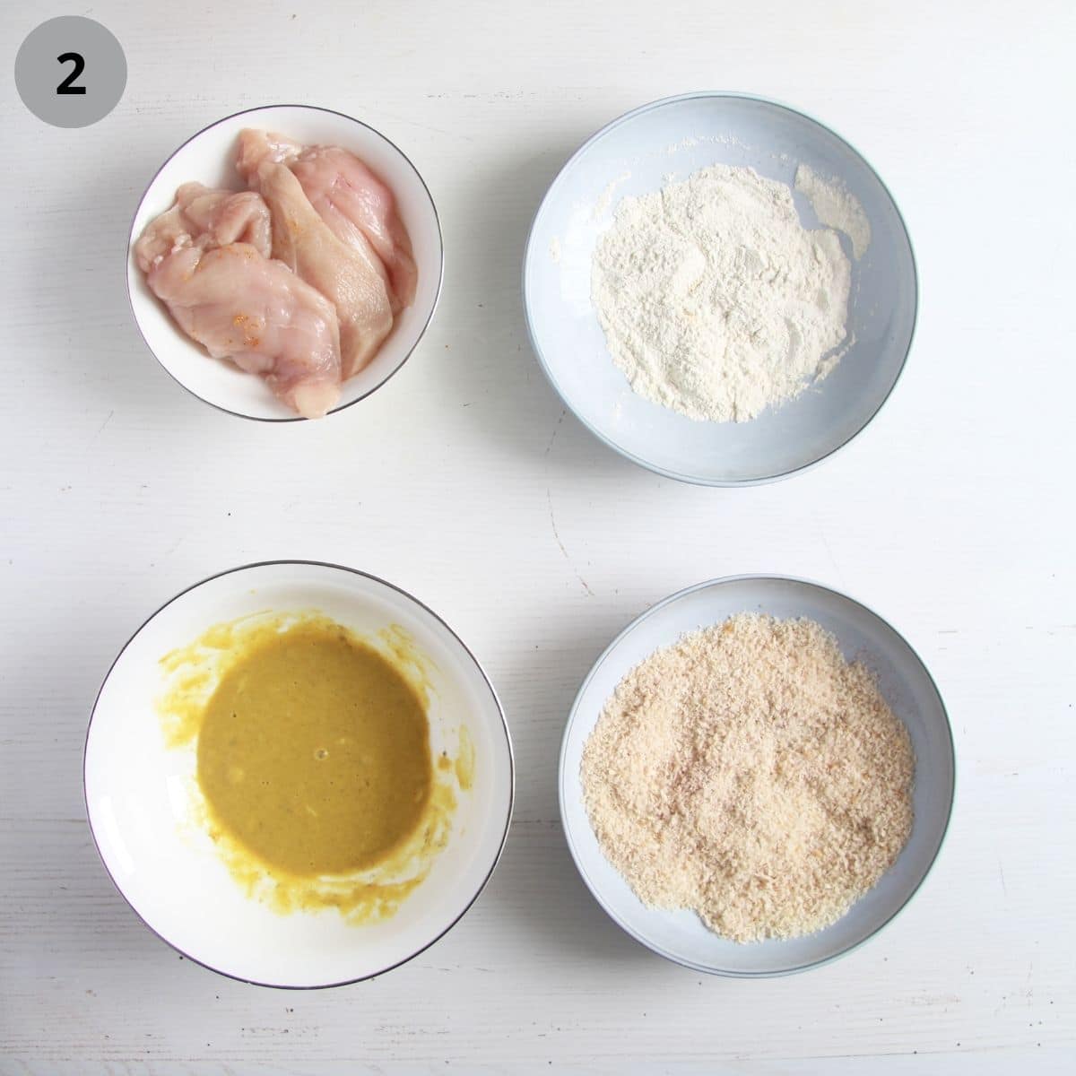 four bowls with raw chicken breast, flour, egg mixture and breadcrumbs.