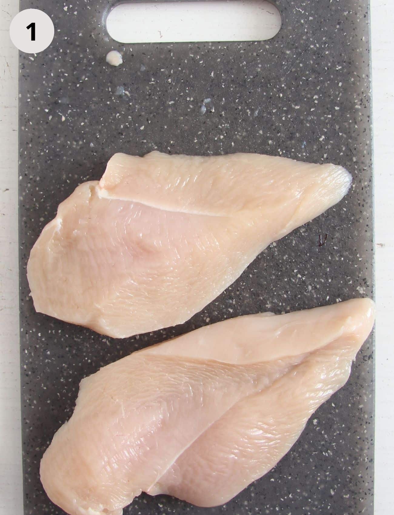 two pieces of raw chicken breast on a dark cutting board.