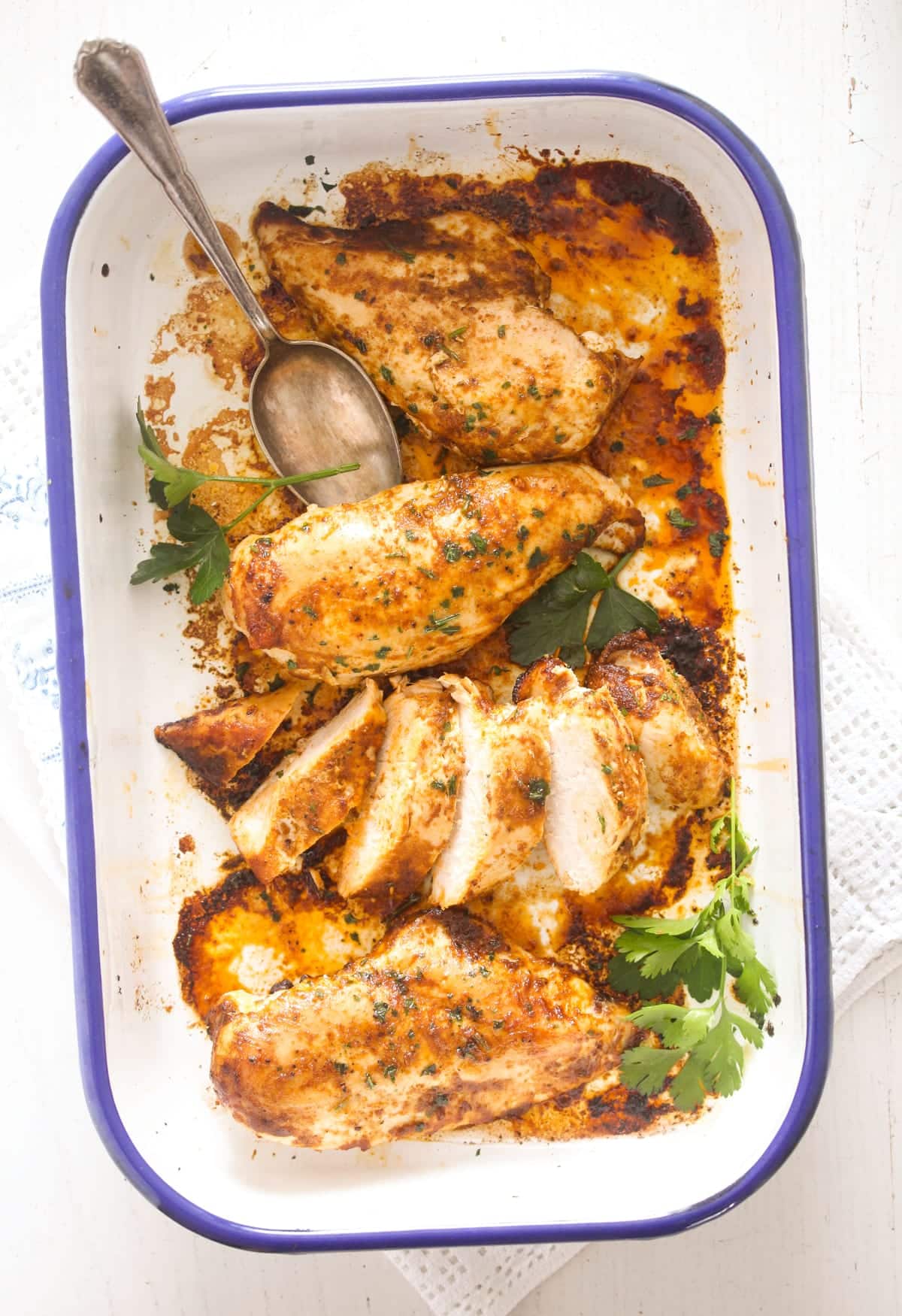 four chicken portions in a white baking dish with blue margins, a spoon in the dish.