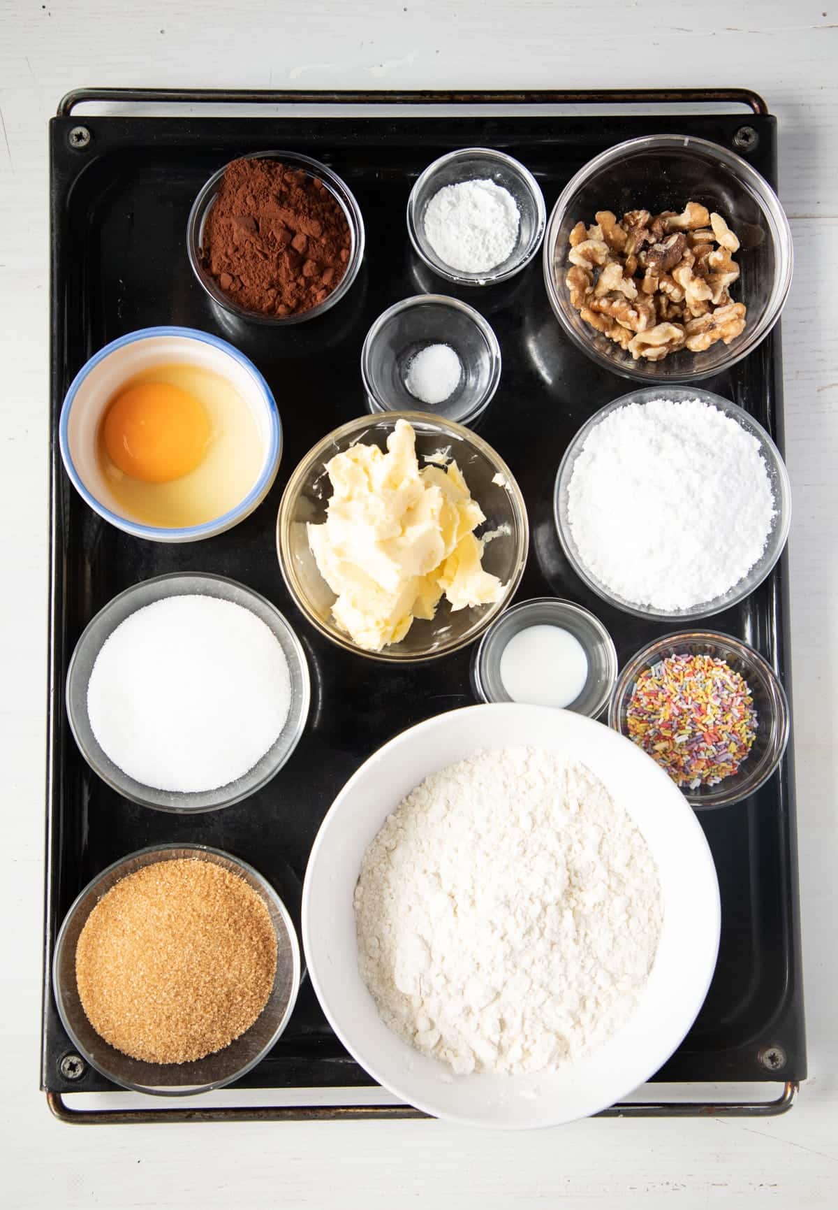 ingredients for making chocolate cookies with walnuts in many small bowls on a baking tray.
