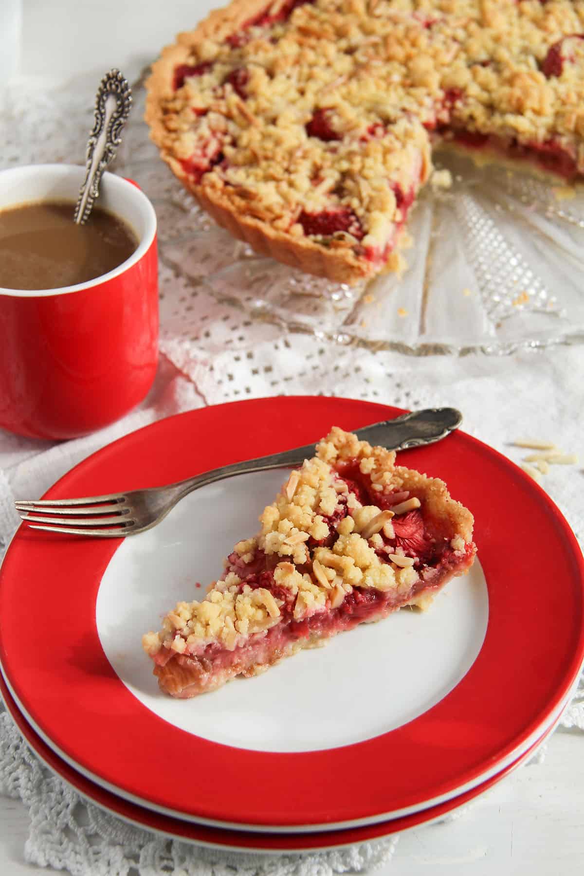 a slice of rhubarb strawberry tart on a plate, a cup of coffee and the rest of the tart behind.