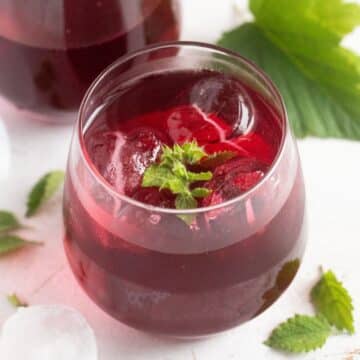 black currant gin served in a glass with ice cubes and lemon balm leaves.