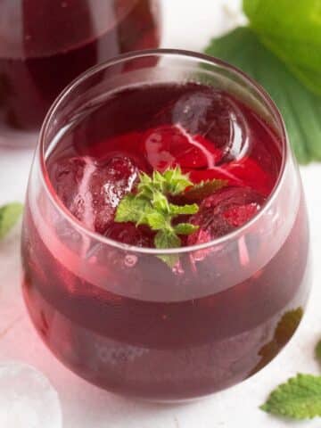 black currant gin served in a glass with ice cubes and lemon balm leaves.