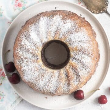 cherry bundt cake sprinkled with icing sugar on a white plate with a few cherries beside it.