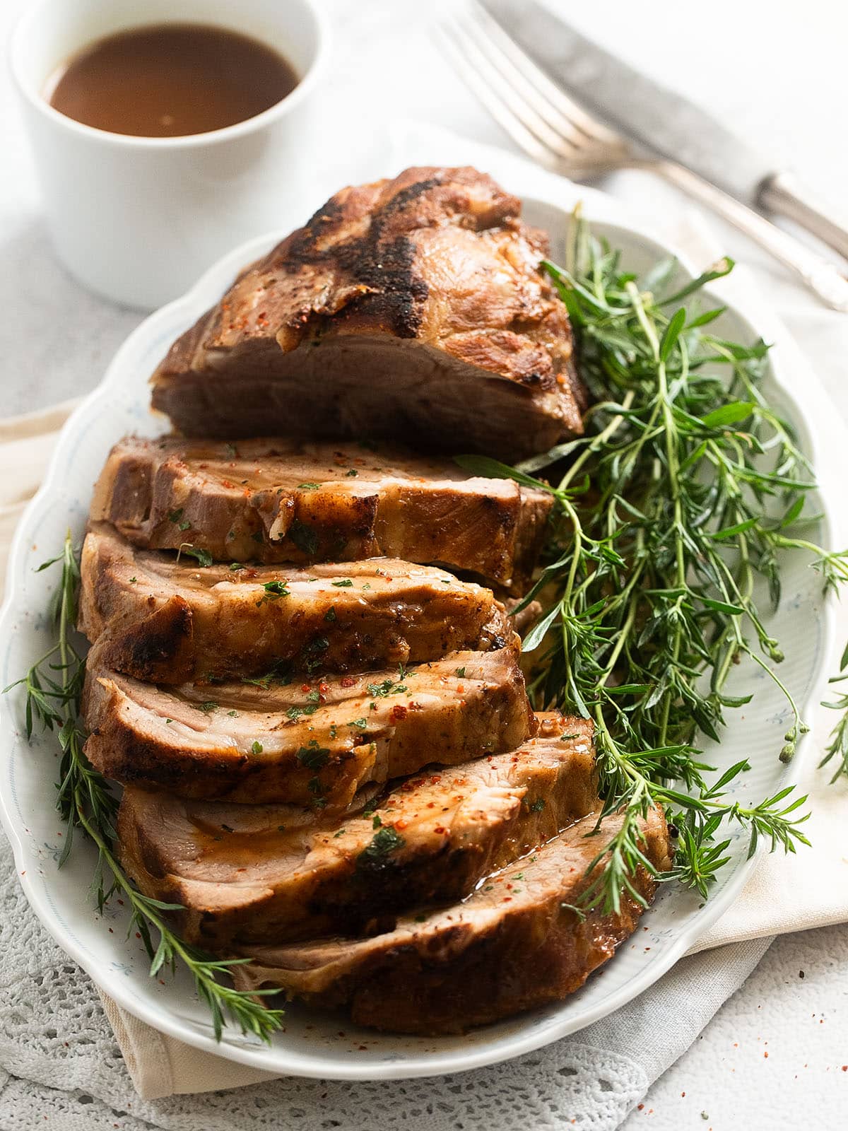 sliced roasted neck of pork on a platter with a bunch of thyme and a small pot of gravy behind it.