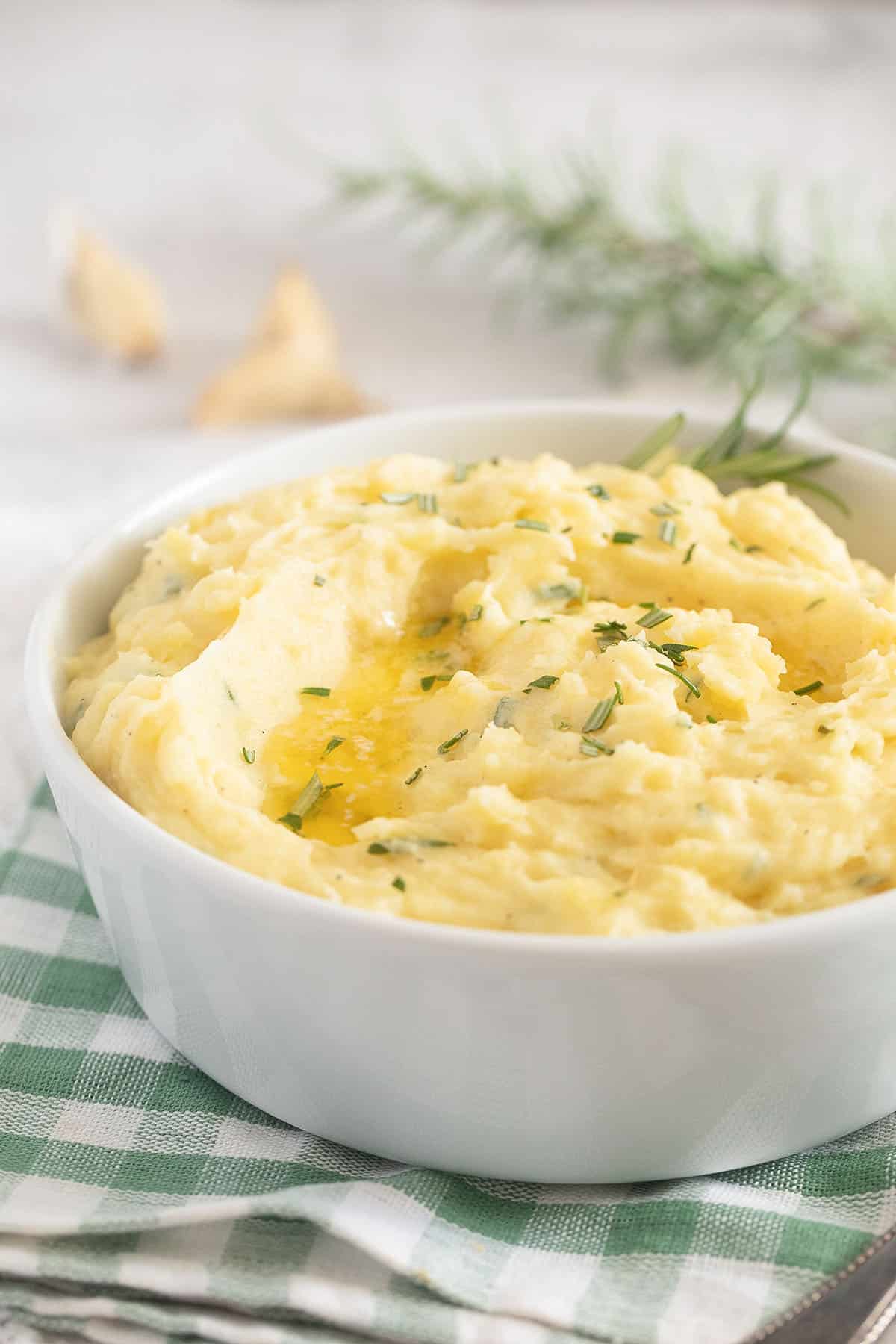 bowl of mashed potatoes with rosemary and garlic on a green and white cloth.
