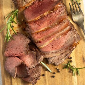 sliced roast beef, rosemary sprigs, and a fork on a wooden cutting board.