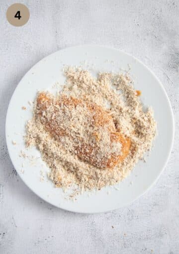 coating one chicken breast with ground almonds on a plate.