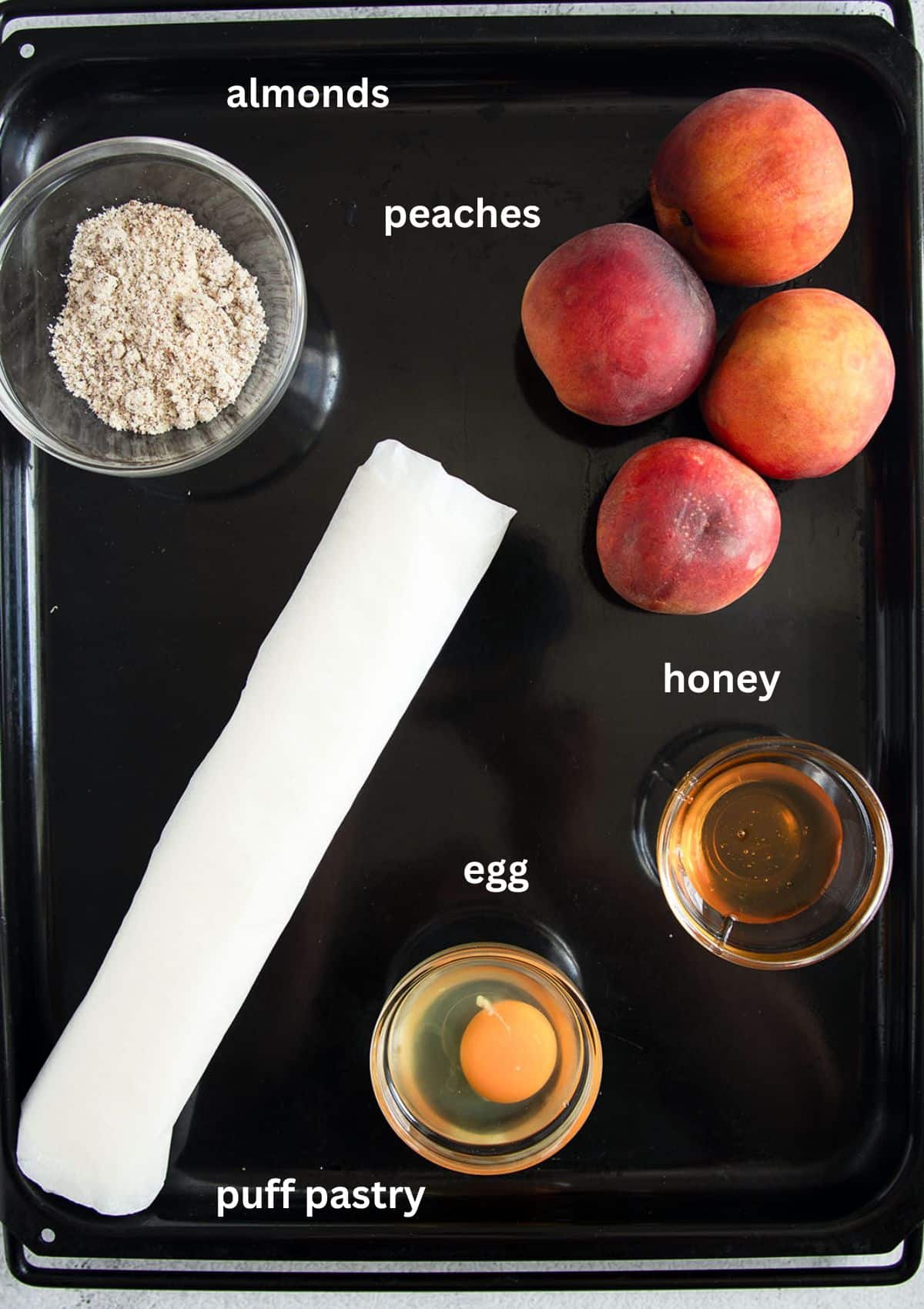 rolled puff pastry sheet, bowl with ground almonds, four peaches, bowls with honey and one egg for making tarts.