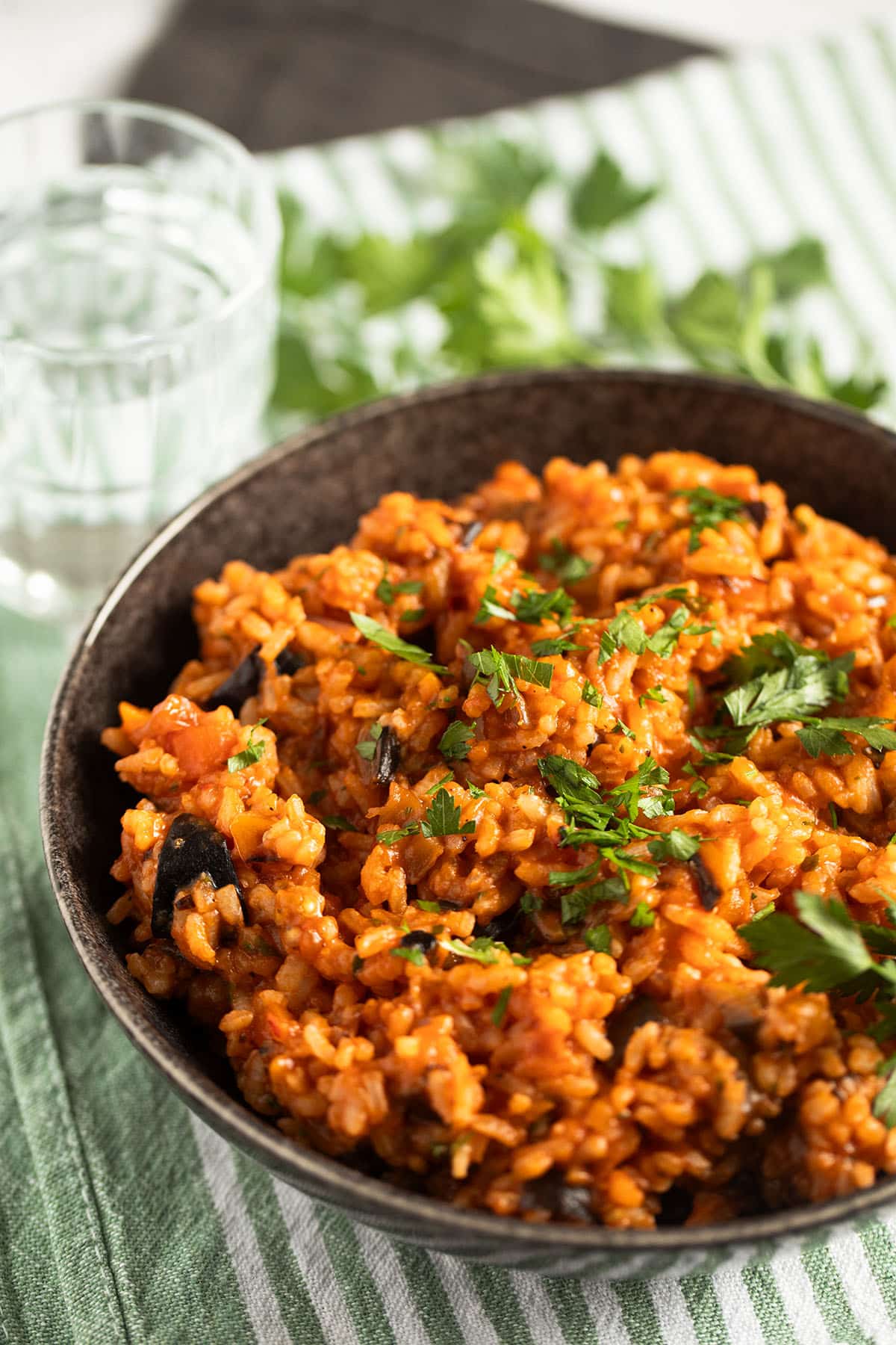 a bowl of rice with eggplants, a glass of water and parsley on the table.